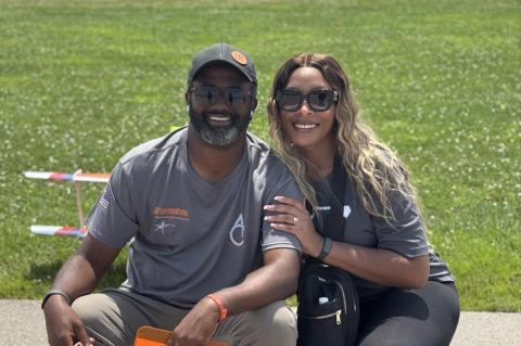 A smiling couple seated on grass, wearing sunglasses and casual clothing.