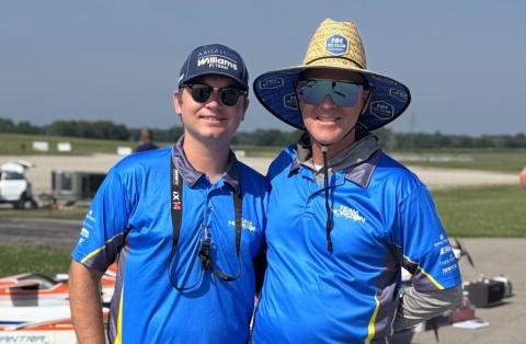Two smiling men in blue shirts and hats at a sunny outdoor event.