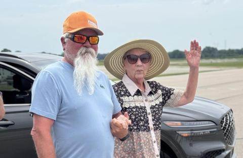 Older woman in sunhat raises hand beside bearded man in sunglasses near a car.