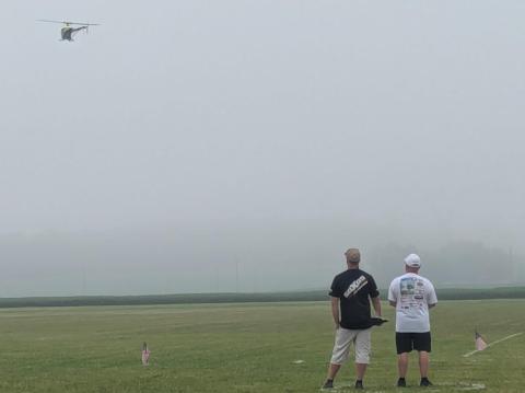 Two men watch a small helicopter fly in a foggy field.