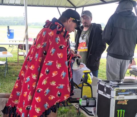 Man wearing a red bird pattern blanket, under a canopy, working with gear.