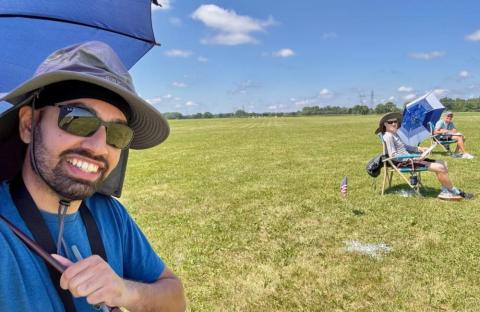 Smiling person with umbrella in a sunny field, others sitting on chairs in the distance.