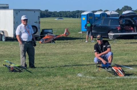 Two men with remote-controlled helicopters on a grassy field.