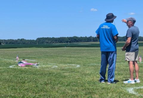 Two people stand in a field, discussing a small model aircraft on the grass.
