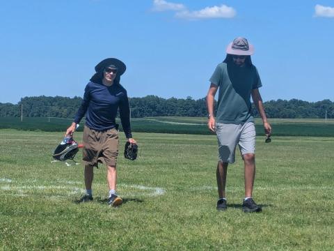 Two men walking on grass, wearing hats; one holds sports equipment.