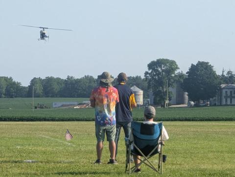 Three people watching a distant flying helicopter over a field.