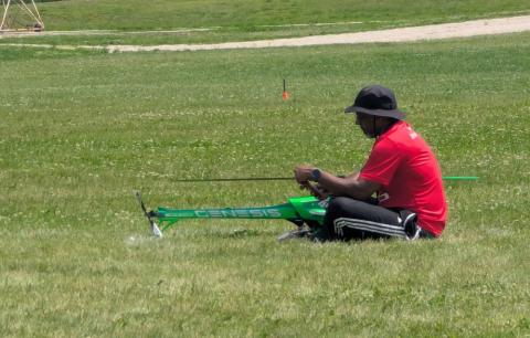 Man in a red shirt sitting on grass, assembling a large green model helicopter.