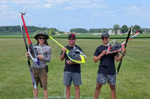 Three people holding model helicopters in a grassy field, smiling.