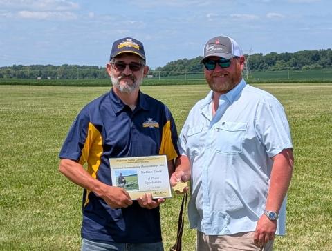 Two men standing in a grassy field, one holding a certificate.