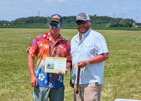 Two men smiling in a field, one holding a certificate, both wearing sunglasses.