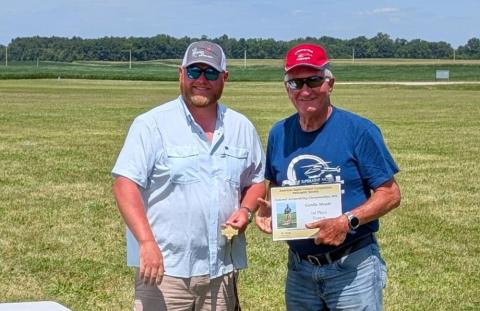Two men standing outdoors, one holding a certificate, both smiling.