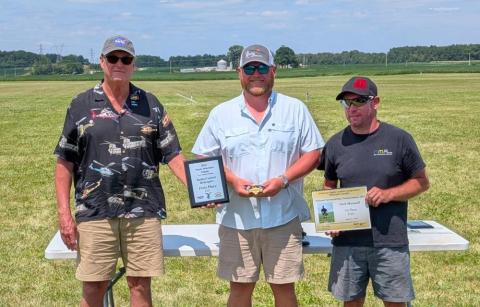 Three men standing on grass, holding certificates under a blue sky.