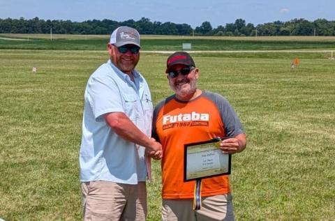 Two men shaking hands outdoors, one holds a certificate, smiling.
