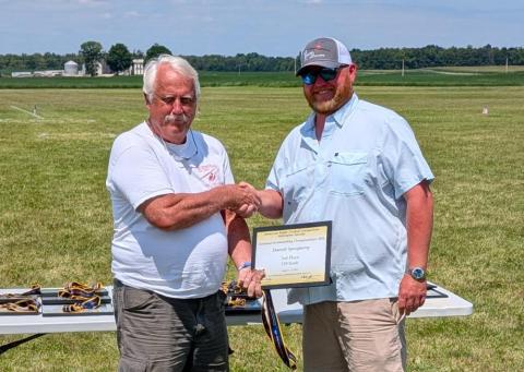 Two men shaking hands, one holding a certificate, outdoors on a grassy field.