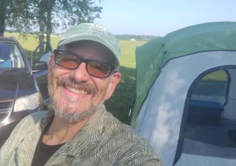Man smiling near a tent and car in a grassy field.
