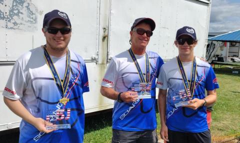 Three men in sports uniforms holding medals and plaques, standing outdoors.