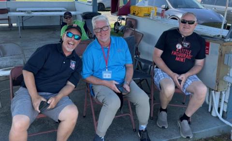 Three men sitting on chairs outdoors, smiling and wearing casual clothing.