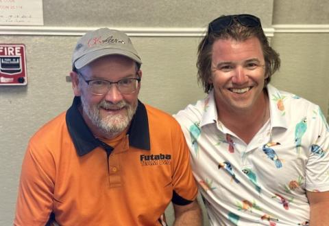 Two smiling people sitting together, one in an orange shirt, the other in a patterned polo.