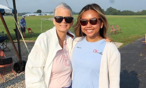 Two women smiling outdoors, wearing sunglasses, with grass and sky in the background.
