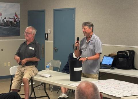 Man speaking into a microphone at a community meeting, others seated nearby.