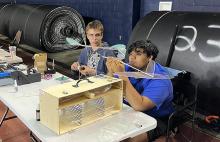 Two people assemble a model aircraft at a table, large rolls in the background.
