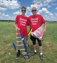 Two people in red shirts holding model planes on grass under a blue sky.