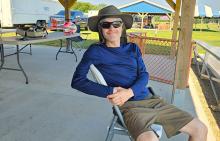 Man in sunglasses and hat, seated outdoors at a picnic area on a sunny day.
