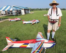 Man with drone controller stands near colorful model planes on grass field.