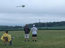 Remote-controlled helicopter flying over a field with three people watching.