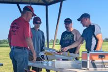 Four men examining a model aircraft on a table outdoors.