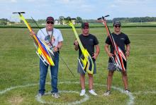 Three men smiling, holding colorful remote control helicopters on grass.